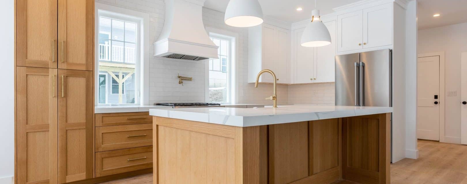 A brightly-lit kitchen with white upper and natural custom wood lower cabinets. There is a large center island with a white quartz countertop. There is custom crown moulding on the ceiling.