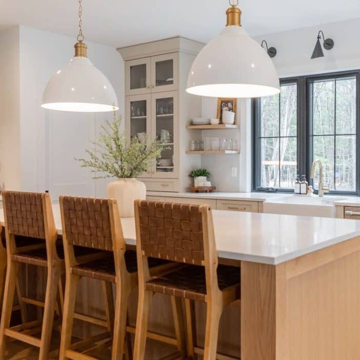 View of a cozy kitchen from behind the large kitchen island. The island is made from neutral hardwood and topped with white quartz. Four wooden bar chairs with woven backs sit at the kitchen island.