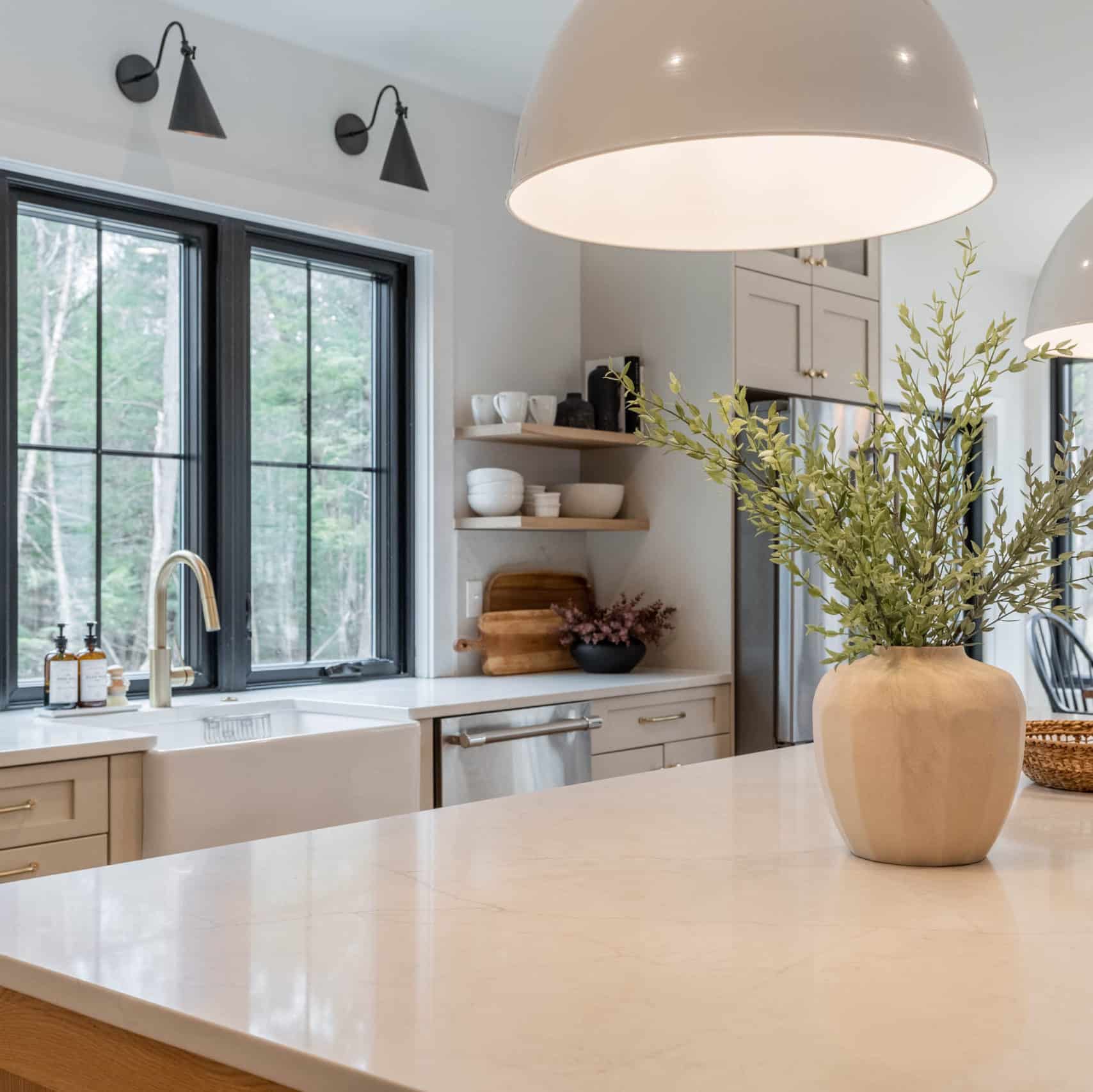 Corner of a cozy, earthy-feeling kitchen. There is a plant with splay leaves in a wooden pot atop a qhite quartz countertop. Floating wooden shelves are stacked with black and white pottery.