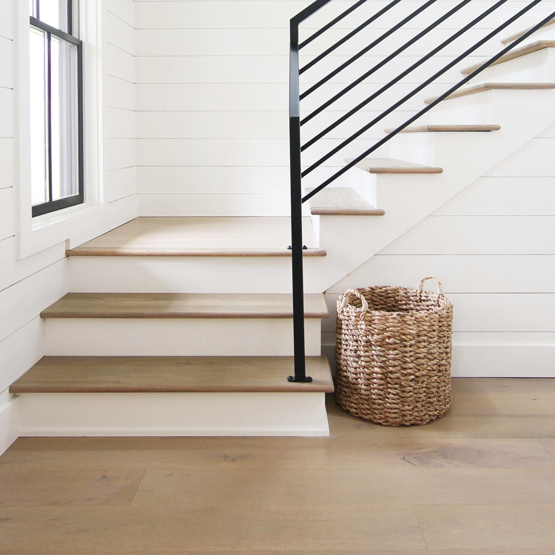 A corner staircase, white with neutral hardwood treads and a black railing. It turns on an angle, not a curve. The walls are white shiplap and a woven basket sits at the foot of the stairs.