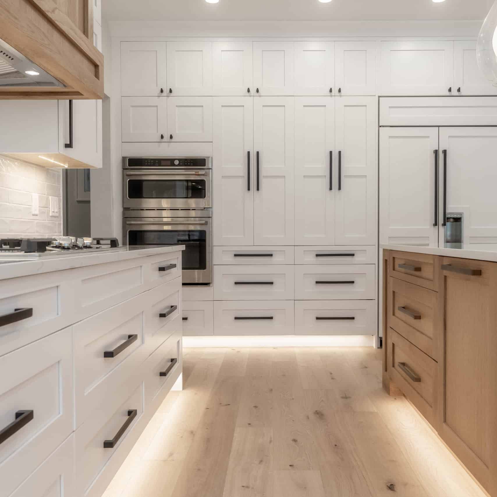 Pantry wall of a large kitchen. The cabinets on the wall are floor to ceiling. They are white shaker cabinets with black hardware. There is a paneled fridge that blends into the cabinets, and two wall ovens.