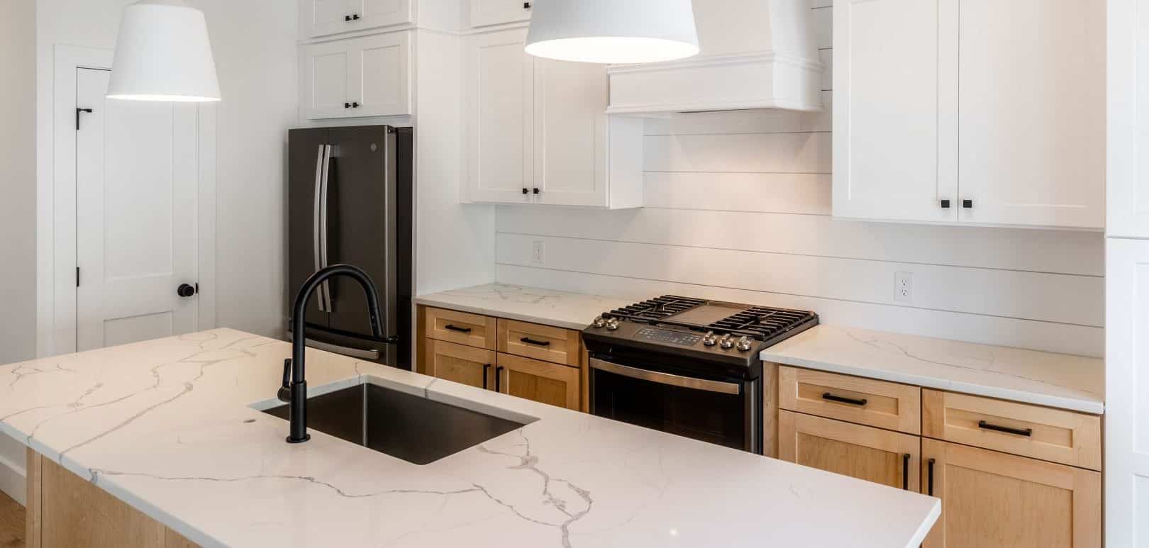 A compact white kitchen with white upper cabinets with black hardware pulls, and lower cabinets finished in a natural maple color. The lower cabinets, as well as the kitchen island across from them, are topped with a marble-like white quartz. The kitchen sink is located on the island, and the oven range against the wall. Behind the oven range is a white shiplap backsplash.