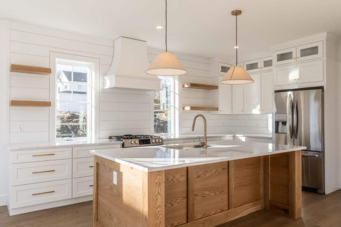 The natural wood base of a large kitchen island stands out against an otherwise all-white kitchen,