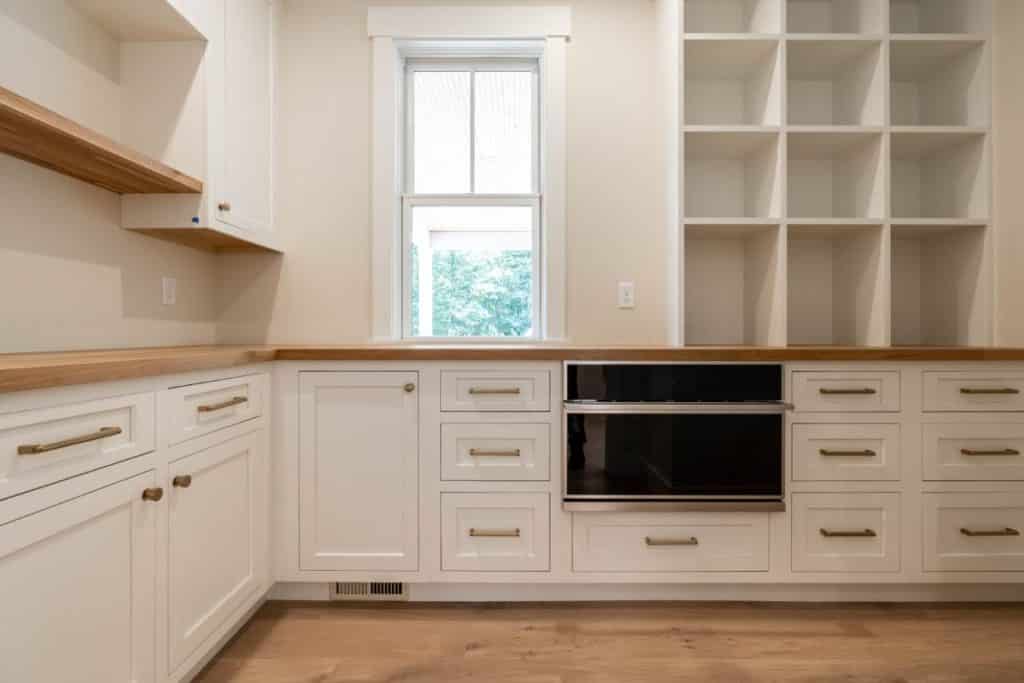The corner of a pantry that is painted mostly white. White cabinets with gold hardware are tucked below a natural wood butcher block countertop. Open shelving sits above the countertop.