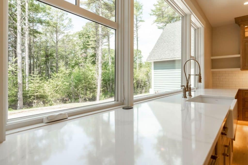 A glance over a white quartz kitchen countertop, through a wide picture window, into a tree-filled backyard.