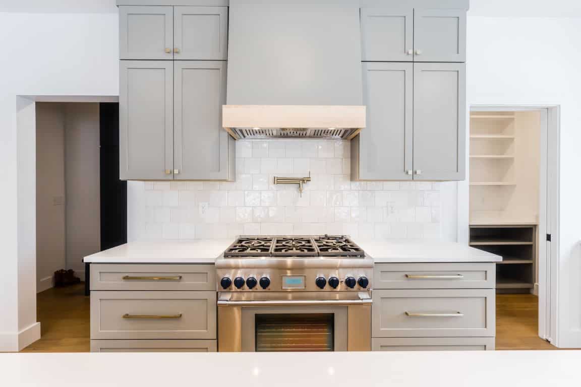 A stainless steel range in front of a backsplash with square, white, textured tiles. Above, a light gray range hood has band of neutral wood at the bottom. The cabinets on either side of the hood are a matching light gray.