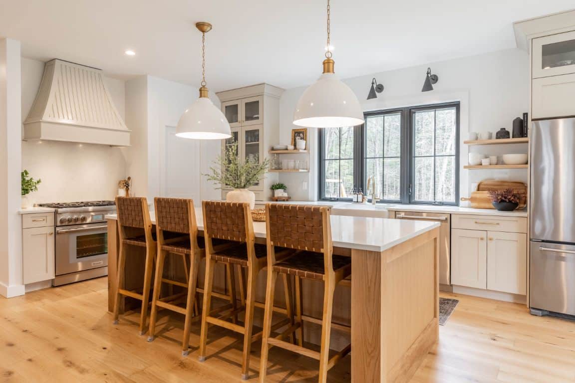 Tall bar chairs with woven backs line up at a wide kitchen island which is in the center of an airy, light-filled kitchen.