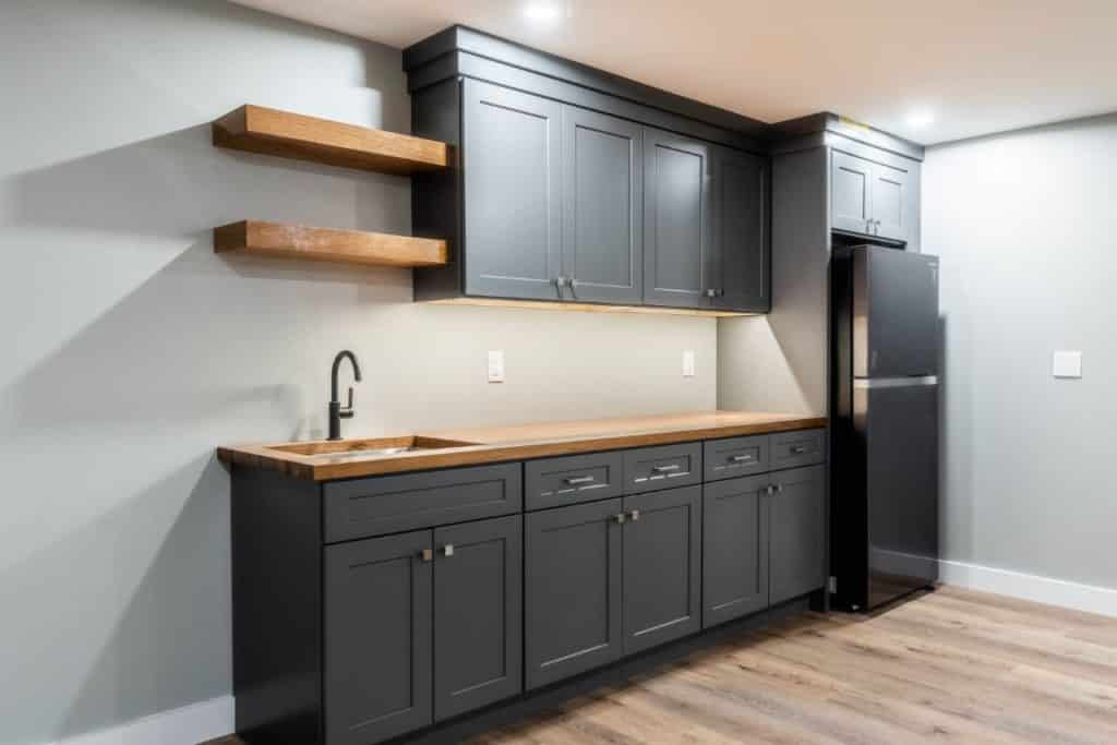 Set of medium gray upper and black lower cabinets against a light gray wall. The lower cabinets are topped with walnut butcher block. There is a small square sink with a black faucet at one end of the lower cabinets. At the other end is a dark stainless steel fridge, ensconced in its own cabinets that have two small doors for storage above.