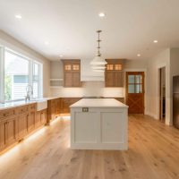 Wide view of a kitchen done in light neutrals. The floor is maple, with natural wood grain. The walnut cabinets are lit from below. In the center of the room is a white island, with white pendants hanging above.